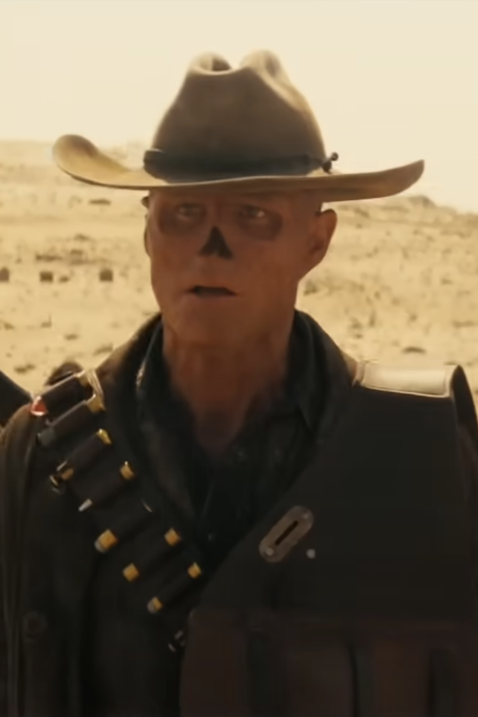 Man with skull face paint in western attire, wearing a cowboy hat and ammo belt, stands in a desert setting