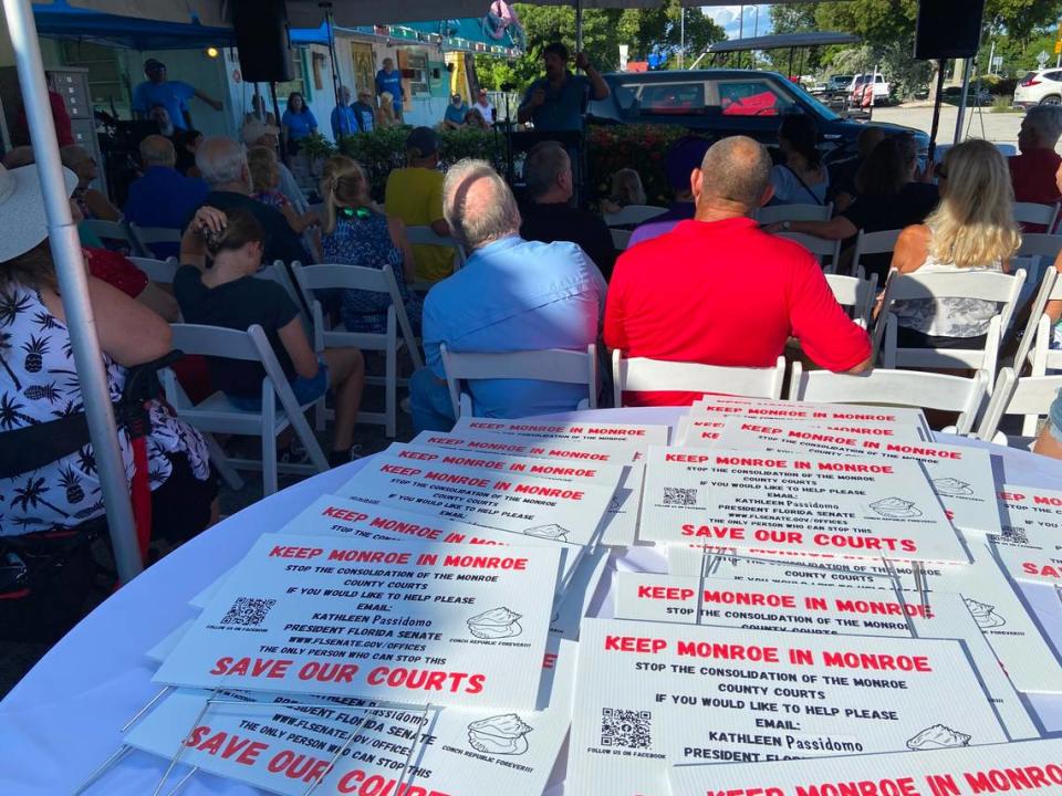 Carteles para jardín se muestran en una mesa detrás de las personas sentadas durante una manifestación en el estacionamiento de un restaurante cubano en Cayo Largo, el domingo 8 de octubre 2023, en protesta contra una propuesta para fusionar los distritos judiciales del estado.