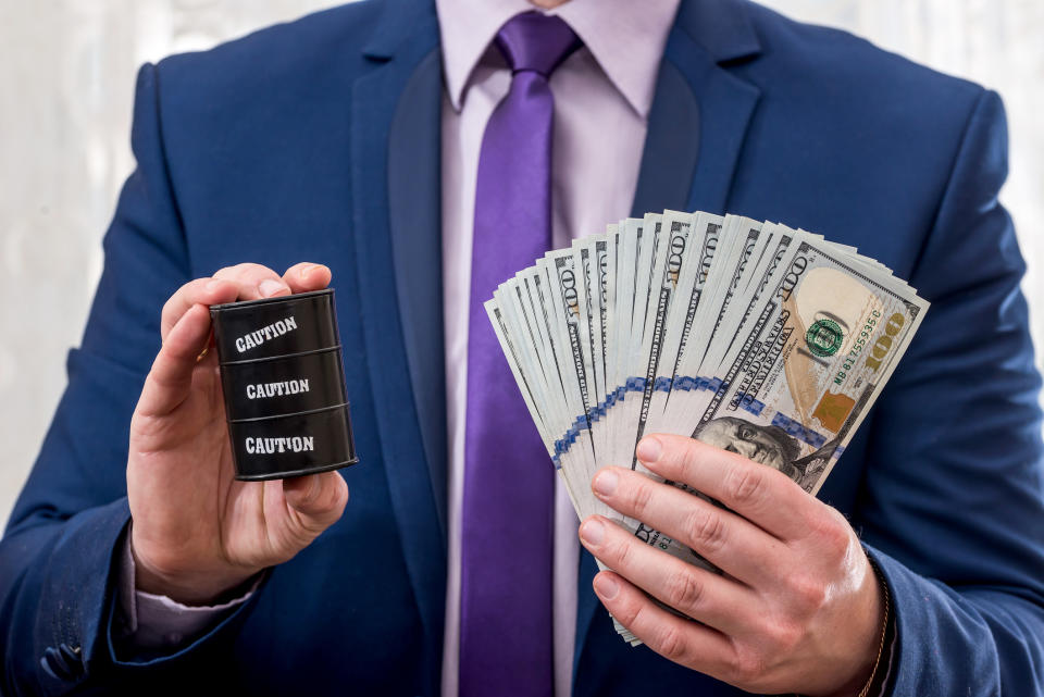 A man holding a miniature black barrel of oil with caution written on it in one hand and fanned-out cash in the other hand.