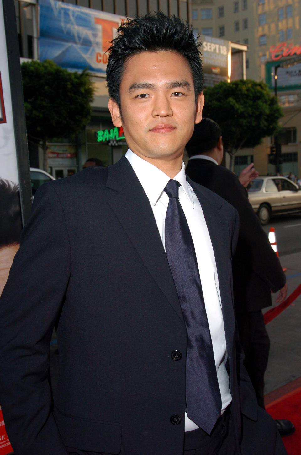 John Cho in a dark suit and tie, posing on a red carpet at an outdoor event