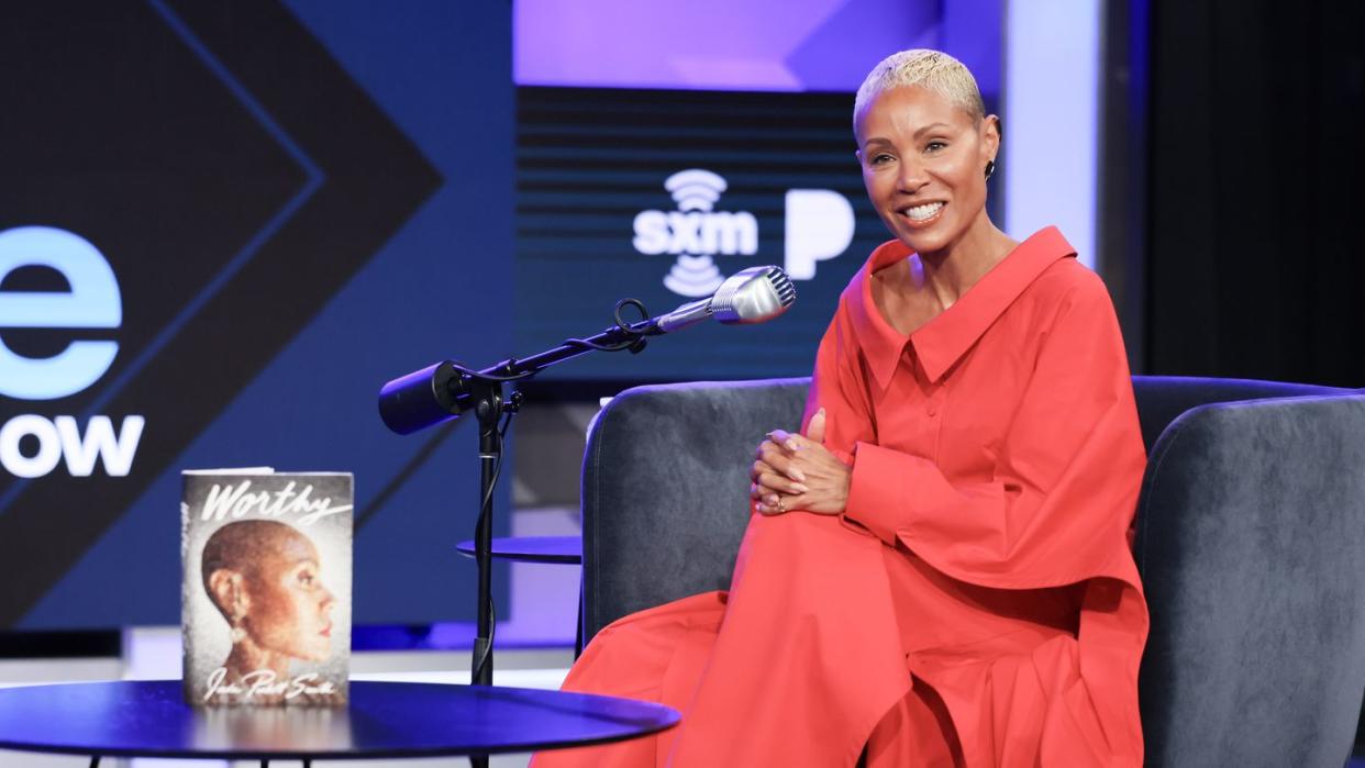 jada pinkett smith smiles and sits in a gray armchair, a microphone is on a stand in front of her as is a round black coffee table with her book worthy standing up, pinkett smith wears a long red dress and bronze high heels