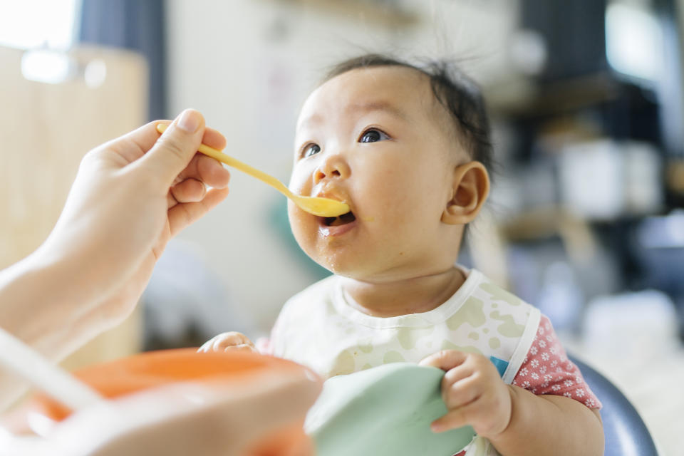 A baby opens his mouth and looks to his mother off-camera as she uses a spoon to feed him