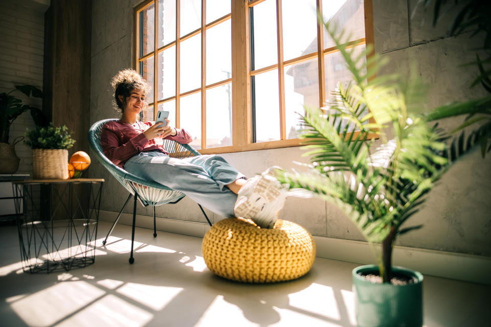 A person relaxes in a chair by a sunny window, looking at their phone, with feet propped on a yellow pouf; plants are in the foreground and background