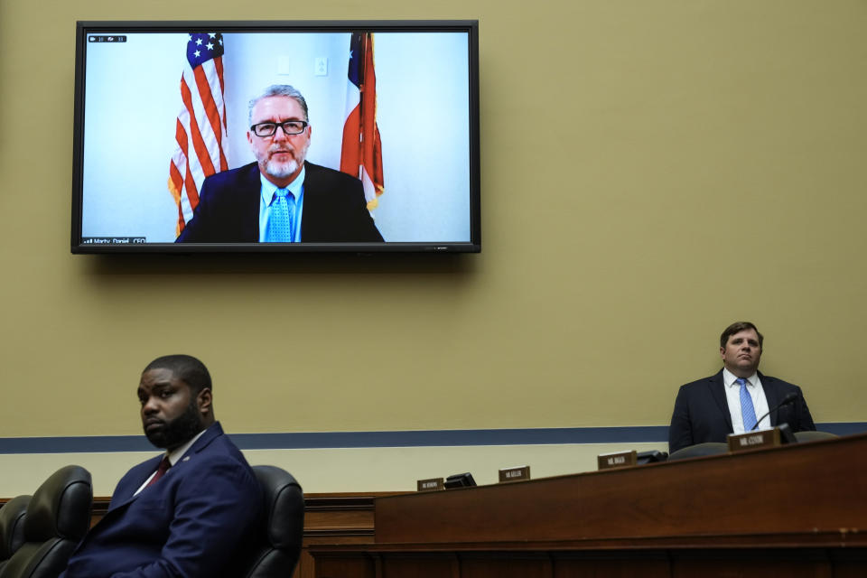 A screen on the wall of a hearing room shows Marty Daniel testifying, as two people listen.