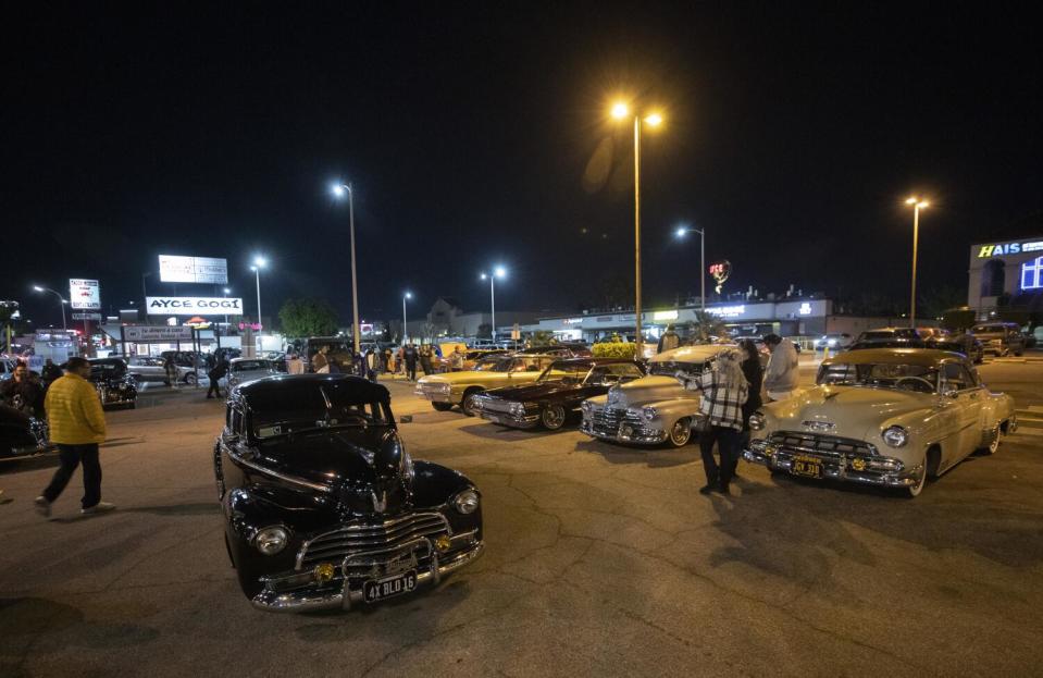 Shiny classic cars are seen at night in a parking lot in the glow of light posts.