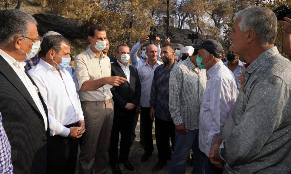 In this photo released Tuesday, Oct. 13, 2020 on the official Facebook page of the Syrian Presidency, Syrian President Bashar Assad, third from left, wearing a mask to help prevent the spread of the coronavirus, speaks with people during his visit to the coastal province of Latakia, Syria. Assad made a rare public visit to Latakia where he toured areas that suffered heavy damage in last week’s deadly wildfires. (Syrian Presidency via Facebook via AP)