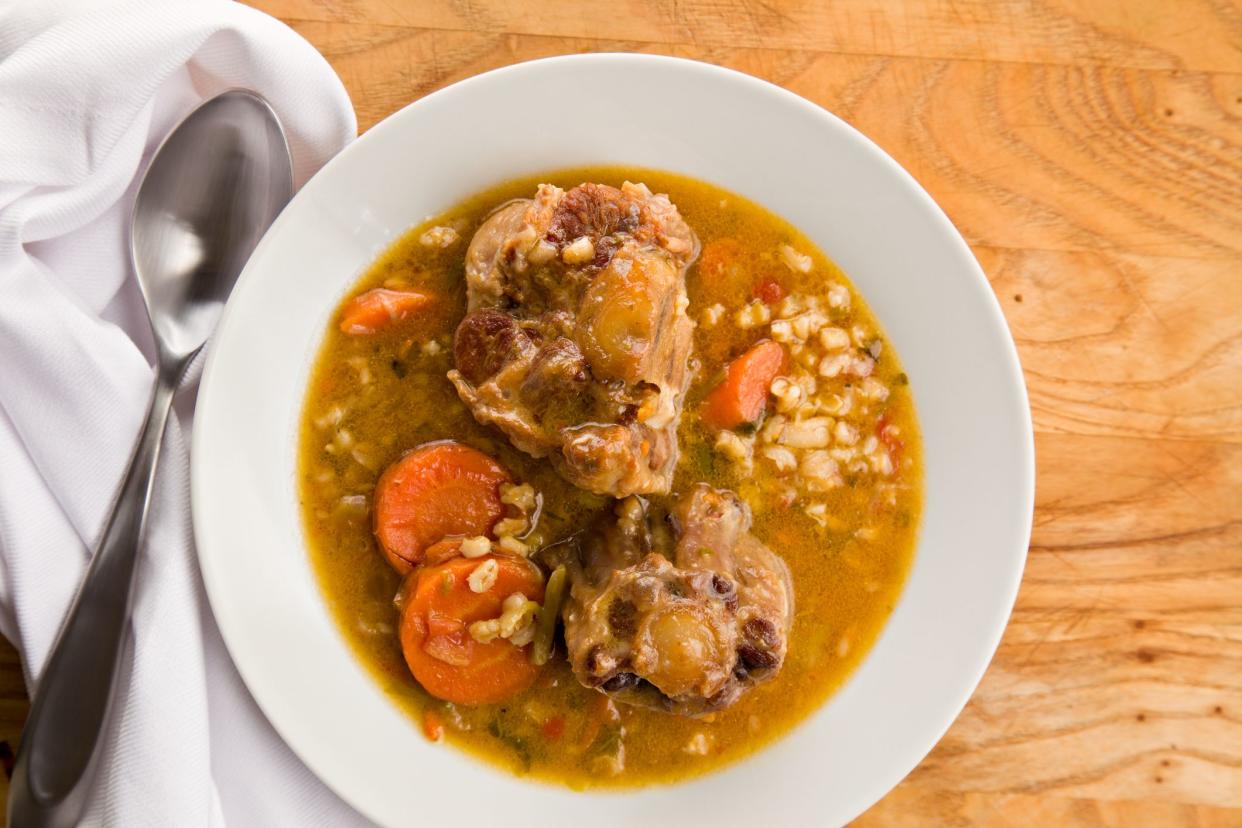 An overhead close up of a bowl of o tail soup a white cloth napkin and  metallic spoon sitting on a wooden surface.