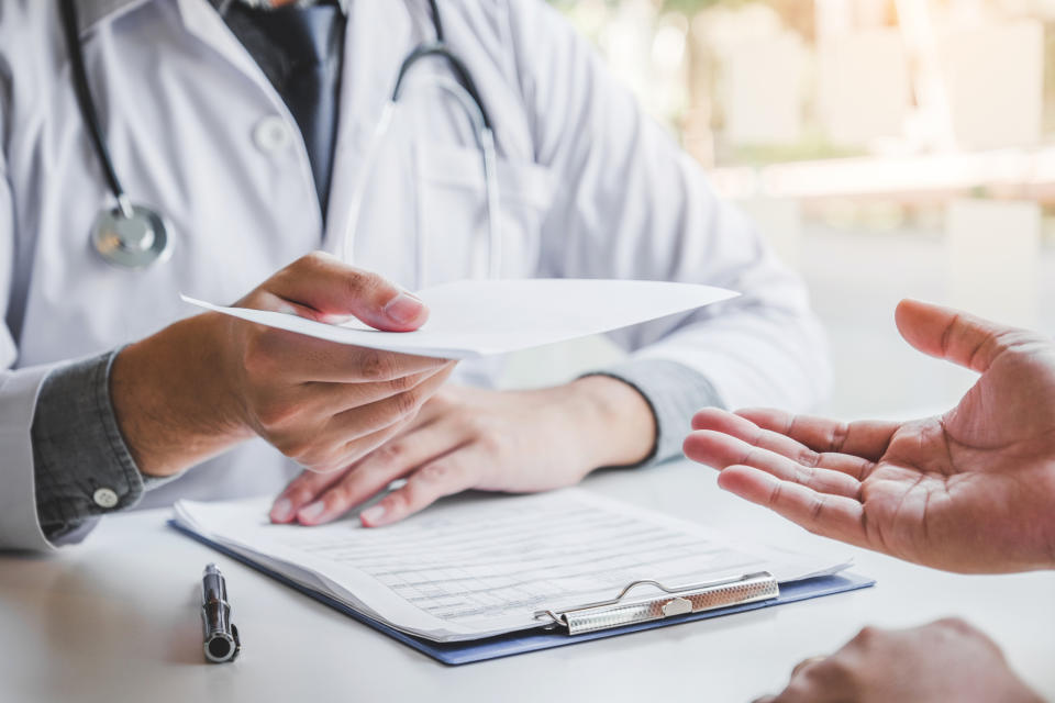 Doctor handing papers to patient across desk, clipboard and stethoscope visible