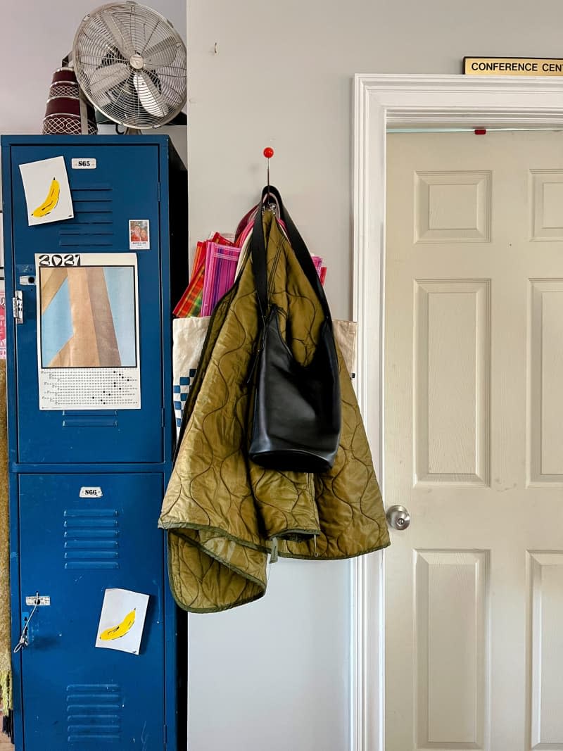 Blue lockers next to a coat hanger with an olive drab jacket and black purse.