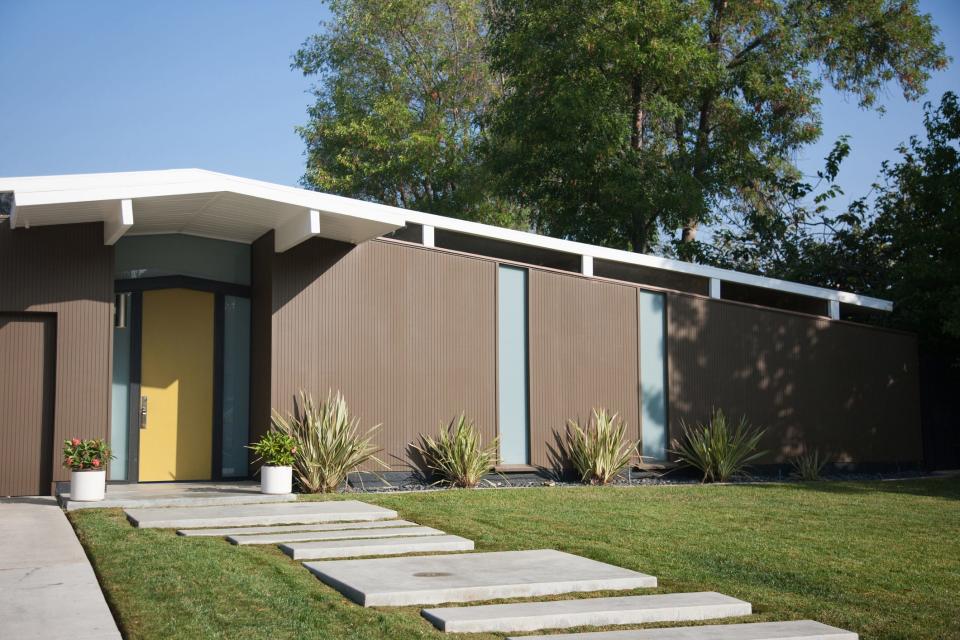 A brown, single-story mid-century modern home with a green door is show. Simple, concrete pavers lined with grass create a walkway to the door.