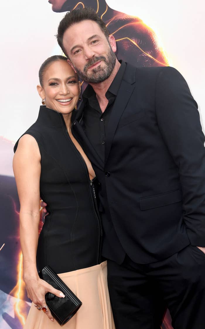 Jennifer Lopez and Ben Affleck pose together on the red carpet. Jennifer wears a sleeveless, high-neck dress with a light-colored bottom, and Ben wears a black suit