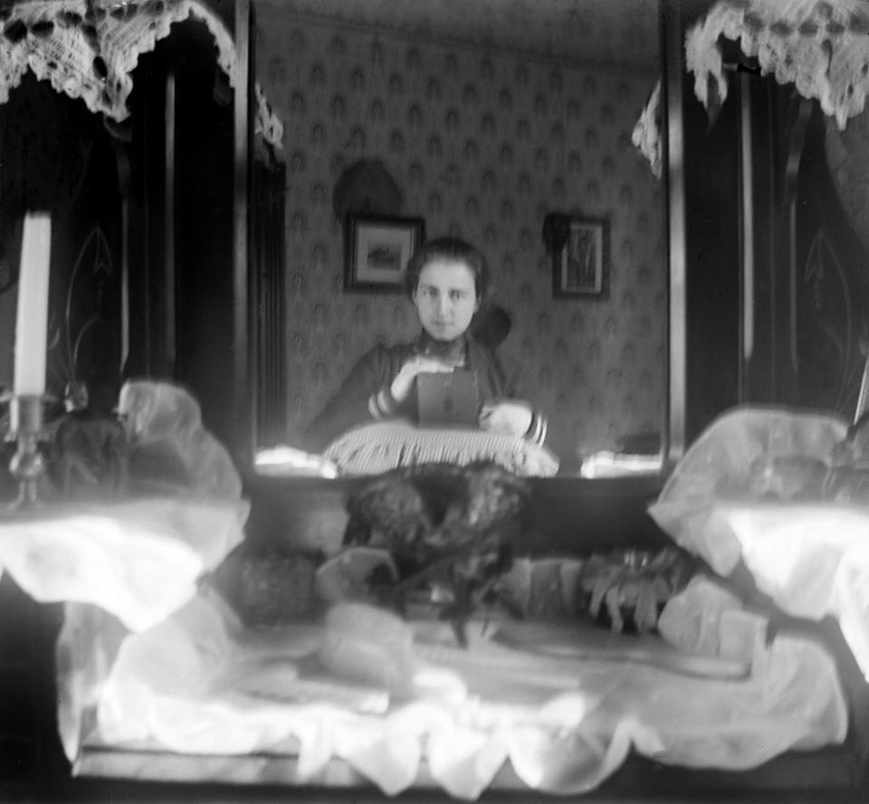 A person, holding an early camera, taking a mirror selfie in a vintage-decorated room. The reflection shows a detailed interior with lace curtains and framed pictures