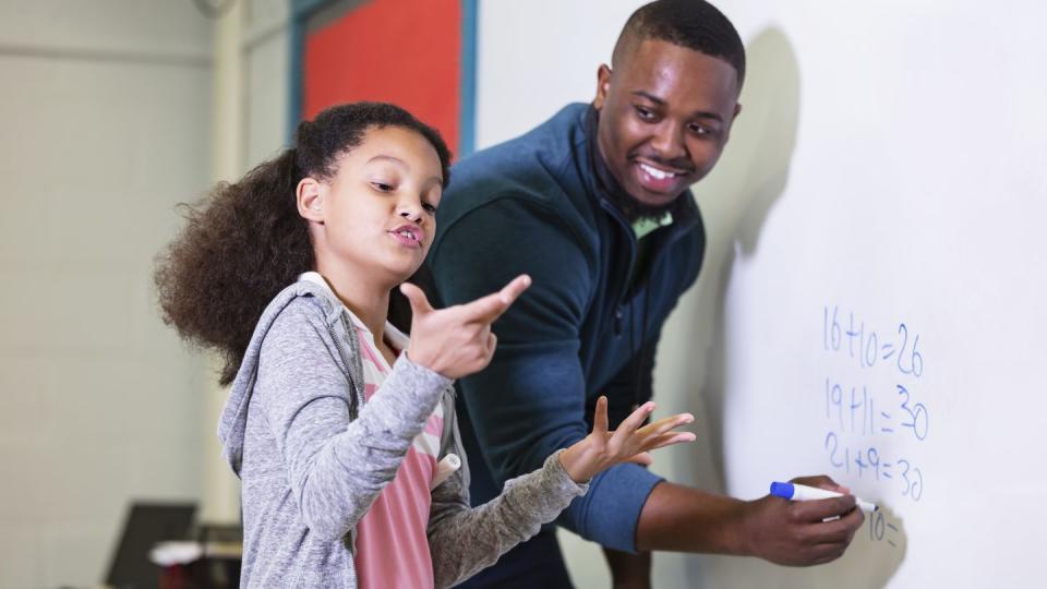 a multiracial 9 year old girl in math class, standing at the whiteboard with her teacher, a young african american man in his 20s the teacher is looking at his elementary student, waiting while she tries to figure out the answer the focus is on the girl