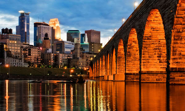 Minneapolis skyline and stone arch bridge at dusk with Mississippi River in foreground.