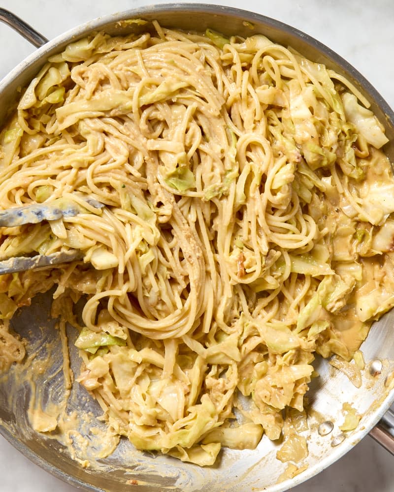 overhead shot of melty cabbage carbonara in a pan, with metal tongs grabbing a bunch of spaghetti.