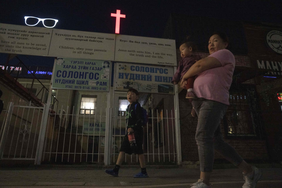 A woman carries a child past a cross and Christian quotes on the street in Ulaanbaatar, Mongolia on Tuesday, Aug. 29, 2023. When Pope Francis travels to Mongolia this week, he will in some ways be completing a mission begun by the 13th century Pope Innocent IV, who dispatched emissaries east to ascertain the intentions of the rapidly expanding Mongol Empire and beseech its leaders to halt the bloodshed and convert. (AP Photo/Ng Han Guan)