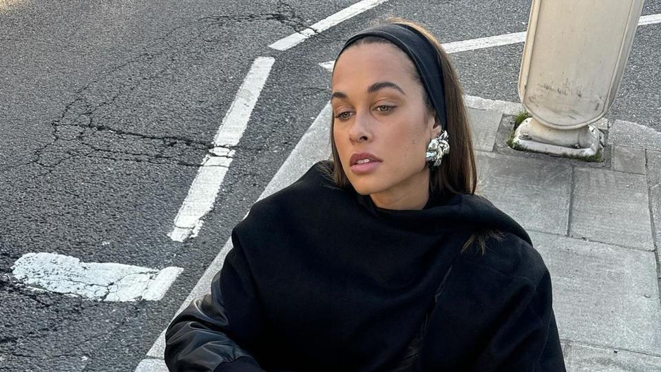 Sarah Lysander wears an all black outfit to sit on a curb in London and accessorises her outfit with a silver metallic Miu Miu bag and bold earrings 