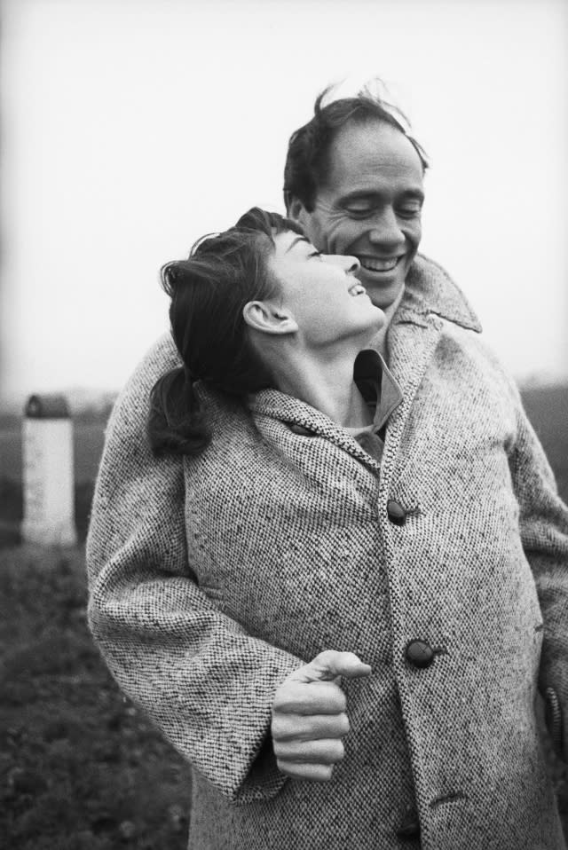 American actor Mel Ferrer (1917 - 2008) buttons up his coat around his wife, actress Audrey Hepburn (1929-1993), on a country road outside Paris, 1956.
