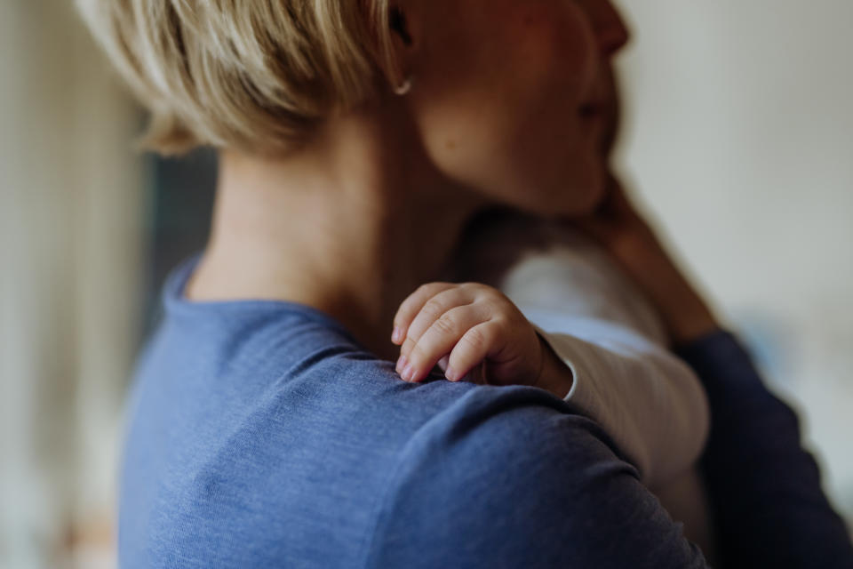 A person with short hair holds a baby close, the baby's tiny hand gently resting on their shoulder