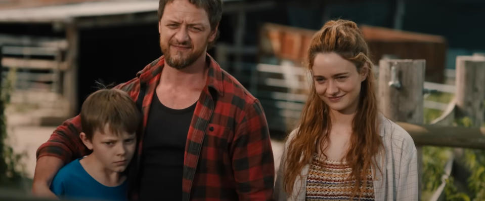 Three people stand outdoors: a man with a beard wearing a plaid shirt, a boy looking serious, and a smiling woman with long hair wearing a cardigan. Names unknown