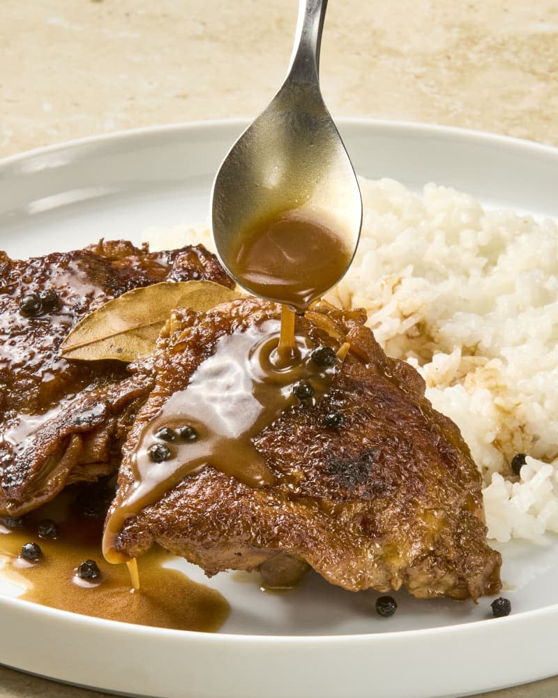 Two chicken thighs prepared in Filipino adobo on a plate with rice on the side with sauce being poured on them.