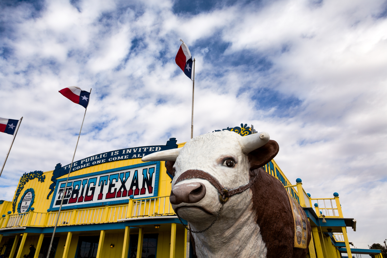 The Big Texan Steak Ranch Restaurant, Amarillo, Texas, cow statue in the foreground with restaurant behind, three texas state flags, a dramatic expansive blue sky with white clouds in the background