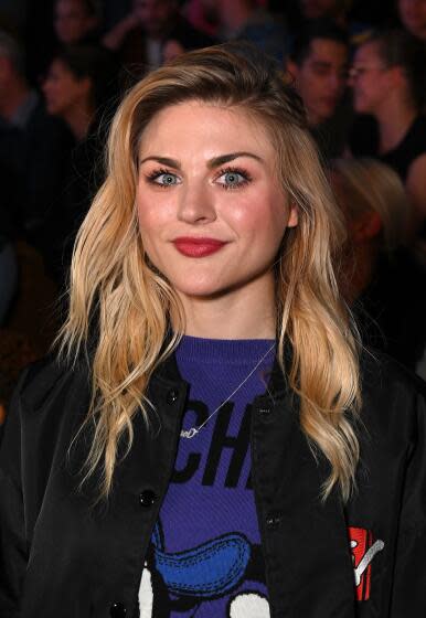 Frances Bean Cobain is smiling and wearing a black jacket and blue t shirt
