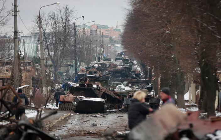 people walking talking on a street full of destroyed and gutted military vehicles