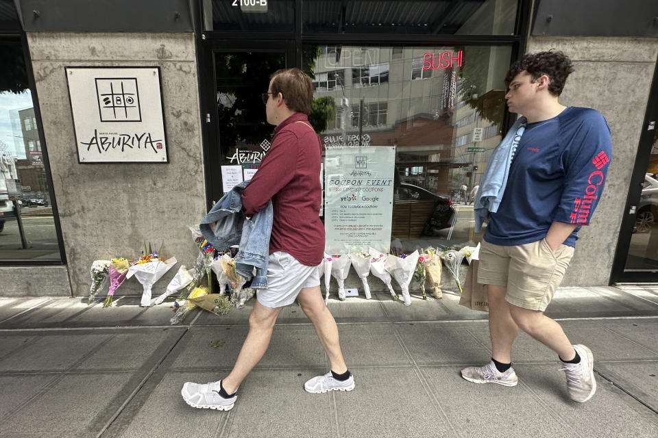 People walk by flowers left at Aburiya, a sushi restaurant that was owned by Eina Kwon and her husband Sung Kwon on Friday, June 16, 2023. Kwon was killed in what appears to have been a random shooting in downtown Seattle this week near the city's famed Pike Place Market. (AP Photo/Manuel Valdes)