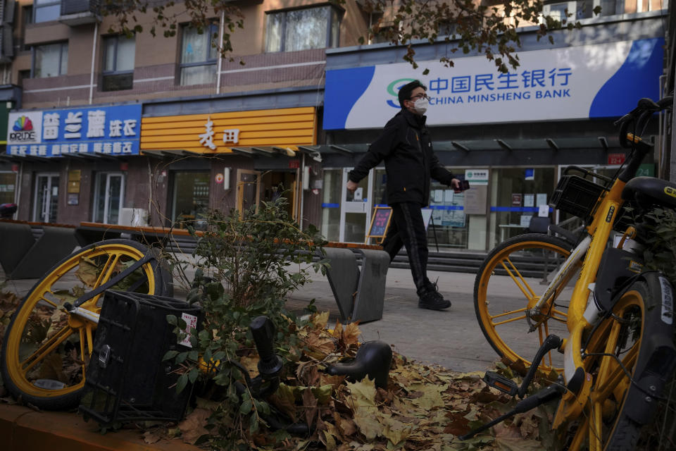 A man walks by a bicycle of bike-sharing company covered with tree leaves near reopened shops after authorities start easing some of the anti-virus controls in Beijing, Wednesday, Dec. 7, 2022. China has announced new measures rolling back COVID-19 restrictions, including limiting lockdowns and testing requirements. (AP Photo/Andy Wong)