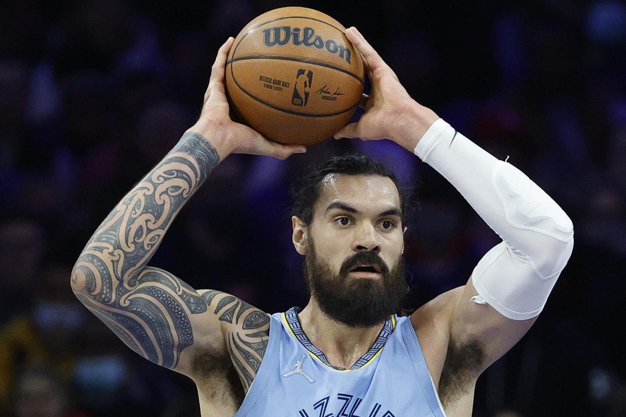 Steven Adams, wearing a Memphis Grizzlies jersey, holds a basketball over his head.