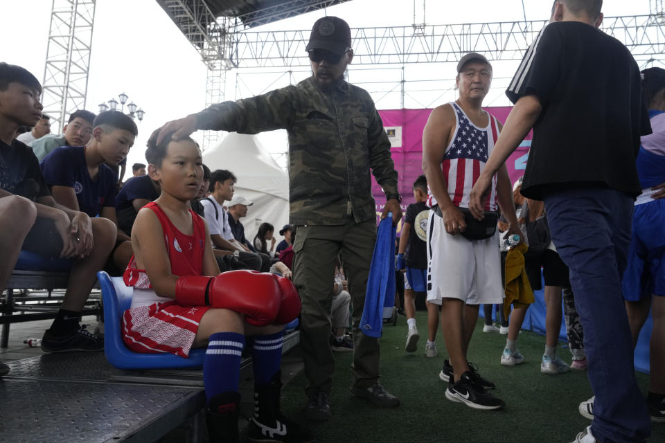 Kherlen Nasantogtokh, center, prepares his twelve-year-old son Gerelt-Od Kherlen, in red, for a bronze medal boxing match on Sukhbaatar Square in Ulaanbaatar, Mongolia, Tuesday, July 2, 2024. Growing up in a Ger district without proper running water, Gerelt-Od fetched water from a nearby kiosk every day for his family. Carrying water and playing ball with his siblings and other children made him strong and resilient. (AP Photo/Ng Han Guan)