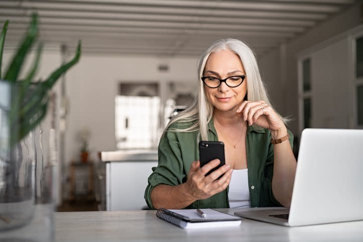 A smiling woman looking at the phone in her hand while sitting at a desk in her living room with her laptop open in front of her.