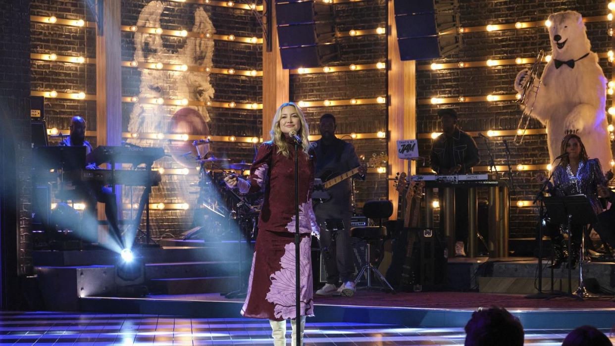 kate hudson stands behind a microphone on a stand on a stage, she wears a maroon and purple dress with white boots, musicians play instruments behind her