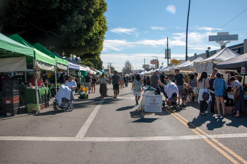 People stroll through a bustling outdoor market with various vendor tents on both sides of a wide walkway