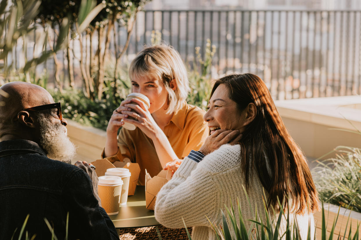 A young woman and a middle-aged woman sit outside at a table, having coffee with an older man.