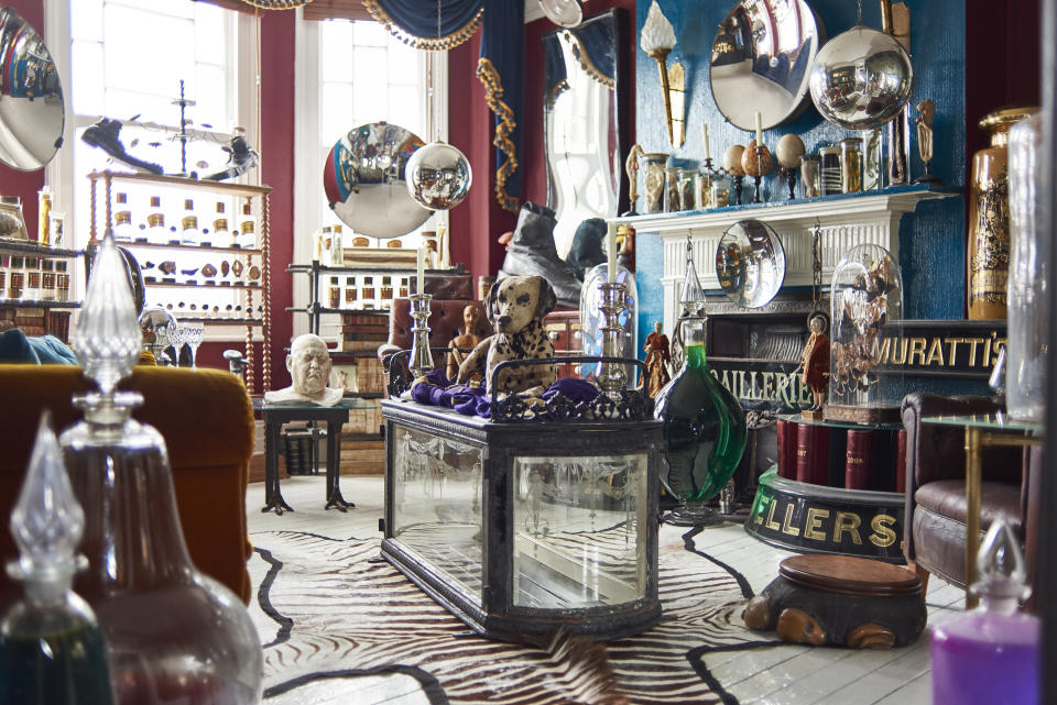 an antique dealer's living room filled with a child's glass coffin carrier, an elephants foot foot stool, a taxidermy dalmatian, a giant leather shoe, decorated ostrich eggs, a plaster death mask, and carboy's filled with coloured liquids