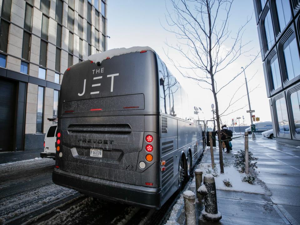 A matte black bus that reads "The Jet" on the side. Passengers with bags are boarding the bus or putting their bags away into the lower storage compartment.A matte black bus that reads "The Jet" on the side. Passengers with bags are boarding the bus or putting their bags away into the lower storage compartment.
