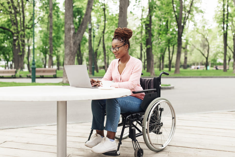 Remote job for people with disability. Cheerful young black woman in wheelchair working online, using laptop at outdoor cafe. African American lady studying or communicating on web