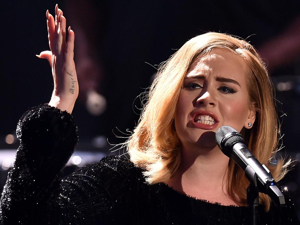 Adele baring her teeth into a microphone with her eyebrows furrowed and hand up in the air.