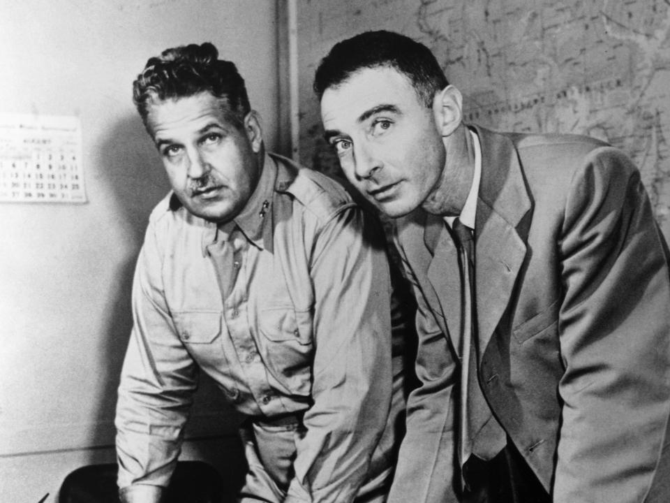 black and white photo of leslie groves in uniform and robert oppenheimer in suit lean on a table in front of a map