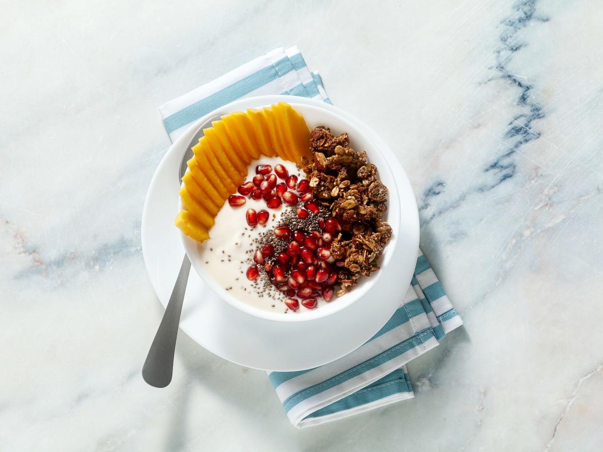 breakfast bowl of granola, yogurt, fresh sweet mango, chia seeds, pomegranate seeds and maple syrup on a blue striped napkin