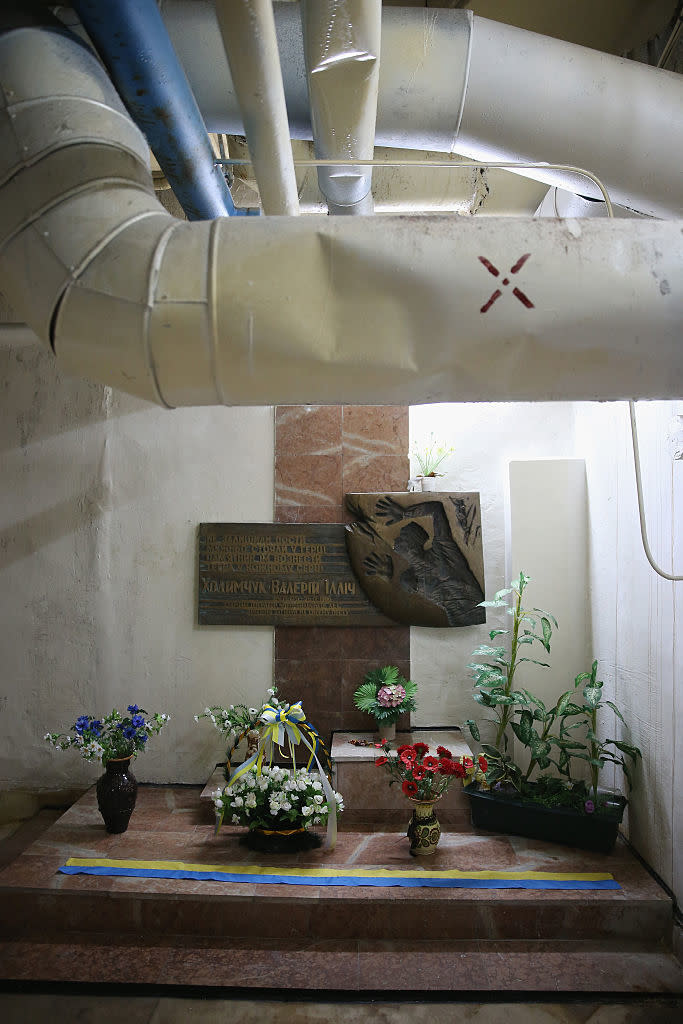 A memorial site with a plaque, flowers, and a cross, surrounded by large pipes above. No people are present