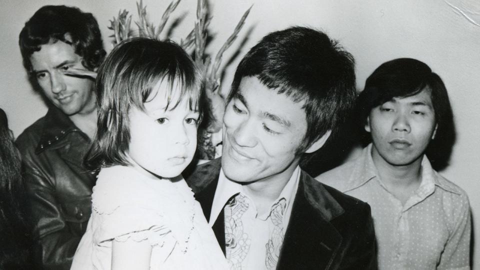 Bruce smiling in black blazer, holding Shannon, at Way of the Dragon premier. Promo. Unknown in background, Bob Wall back left, partially obscured. Wrinkle damage to photo.