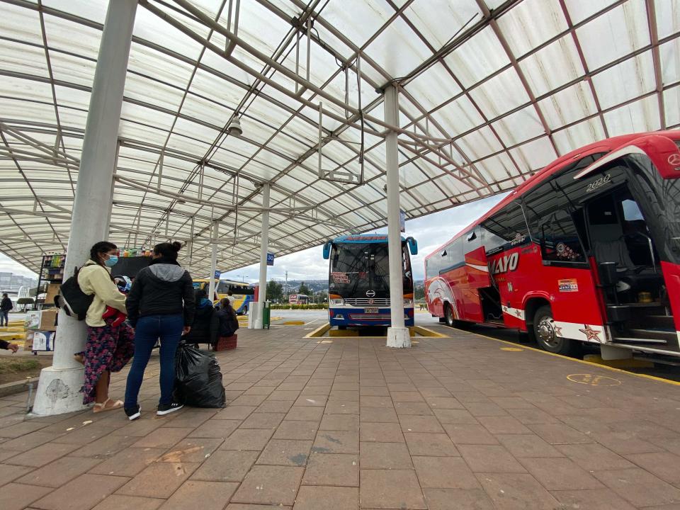 People waiting in a bus terminal with two large buses parked.