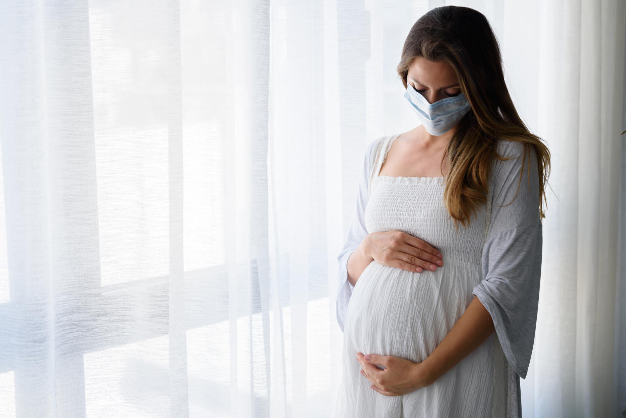 Pregnant woman standing by the big window with face medical mask on. Worries about child birth during pandemic.