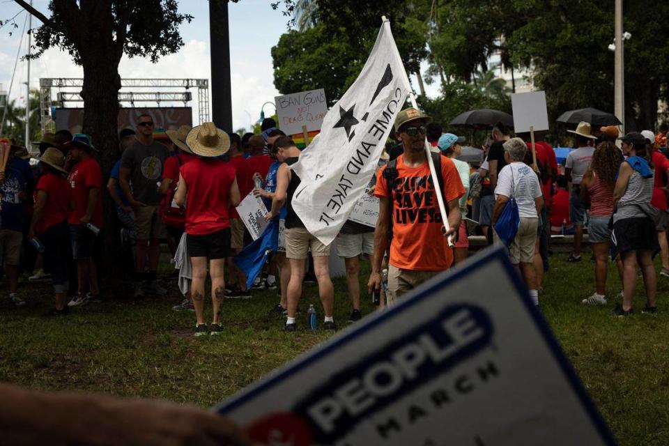 Um homem aparece com uma bandeira “'Venha e pegue' com uma arma automática em resposta à Marcha Anti-Ódio liderada por Nós, o Povo, no domingo, 2 de julho de 2023, no centro de Fort Lauderdale.  Um manifestante ao fundo carregava uma placa que dizia: “Proibir armas, não drag queens”.