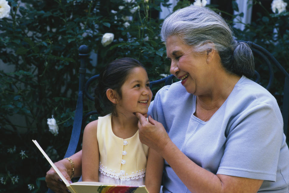 An elderly woman with gray hair and a blue top sits on a bench, smiling and gently touching the chin of a young girl in a sleeveless dress holding a book