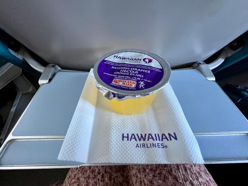 Tray pulled out at writer's airplane seat with a cup of pineapple and a Hawaiian Airlines napkin on tray