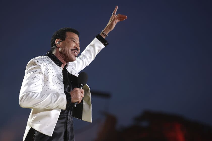 Lionel Richie smiles, holds a microphone and points toward the sky while wearing a white, bedazzled jacket and black pants
